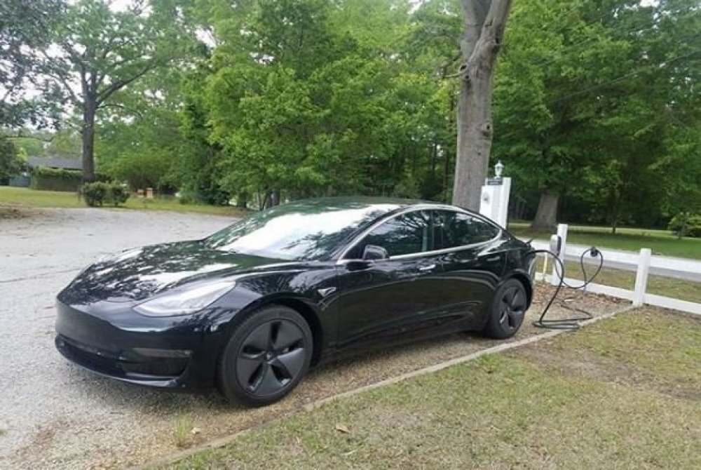 A black electric car is parked outdoors next to a white charging station at Abingdon Manor, surrounded by trees and greenery. The car is plugged in and appears to be charging, reflecting the convenience of EV charging at Abingdon Manor.