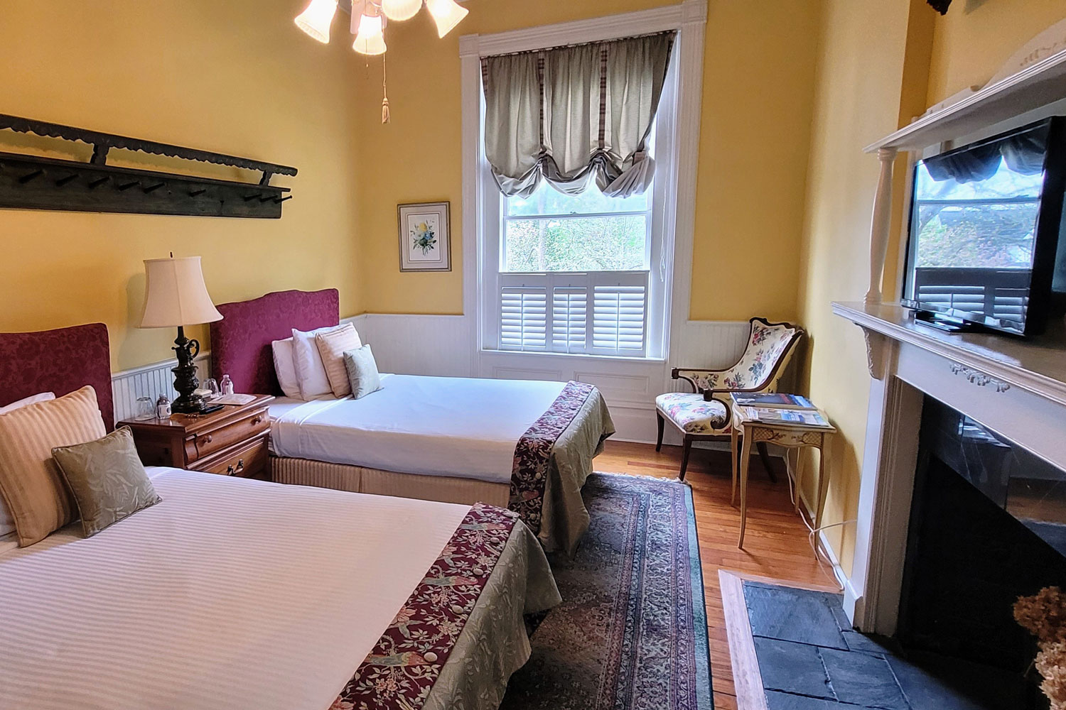 A cozy bedroom with two double beds, floral-patterned bedding, a window with striped curtains, a small chair, a nightstand with a lamp, and a TV above a white fireplace offers charming lodging in Florence SC.