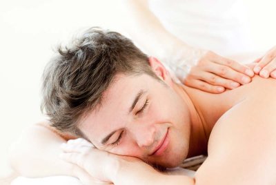 A man with dark hair lies face down, smiling and relaxing with his eyes closed while receiving a shoulder massage from a person whose hands are visible, enjoying the soothing touch provided by massage services near Florence SC.