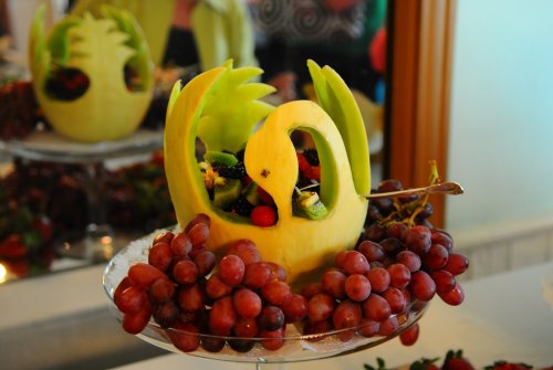 A fruit display, perfect for intimate weddings in South Carolina, features a swan carved from melon, filled with mixed fruit pieces, and surrounded by clusters of red grapes on a glass stand. A mirror in the background reflects the arrangement.