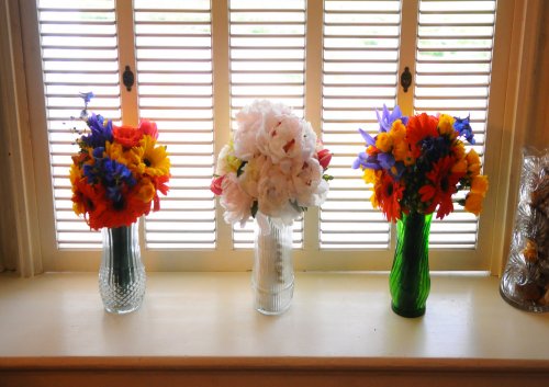 Three vases with colorful flower bouquets sit on a windowsill in front of white shutters. Reminiscent of intimate weddings in South Carolina, the outer vases hold bright mixed flowers, while the center vase displays elegant white blooms.