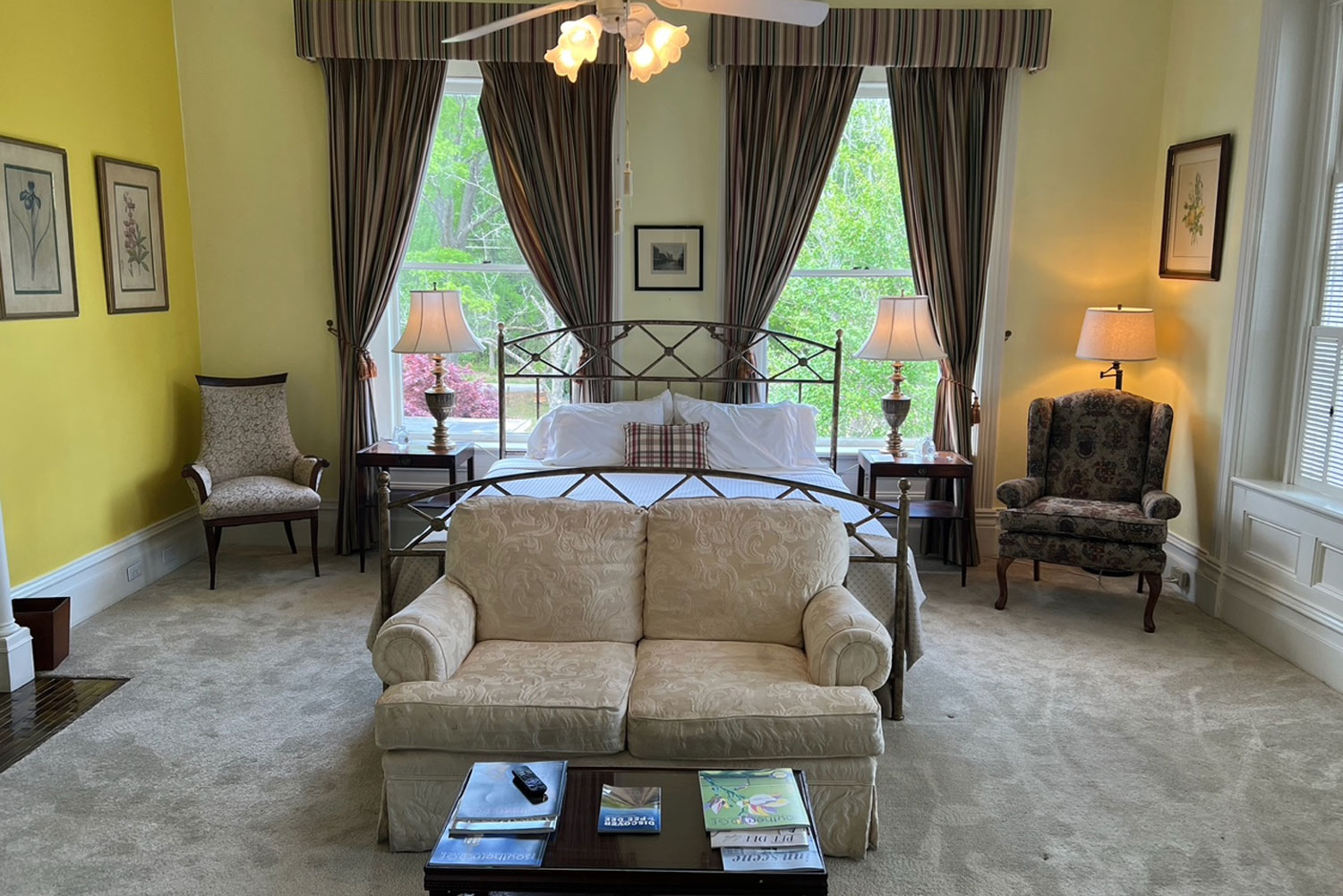 A bright bedroom, perfect for lodging in Florence SC, features a large bed, two armchairs, two lamps, and a cream sofa at the foot of the bed. Large windows with striped curtains let in natural light, creating an inviting and relaxing atmosphere.
