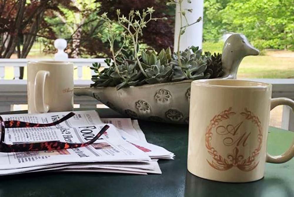 Two coffee mugs, a face mask, newspapers, and a ceramic planter with succulents sit on a green table on a porch with trees and a white railing in the background—a perfect spot to relax while planning your day and looking up things to do in Latta and Florence SC.