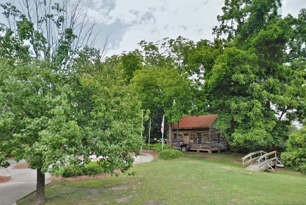 A small wooden cabin with a rusty red roof sits among green trees, near a wooden footbridge and a flagpole, on a grassy lawn under a partly cloudy sky—a peaceful scene reminiscent of the quieter charms you might discover while exploring things to do in Latta and Florence SC.