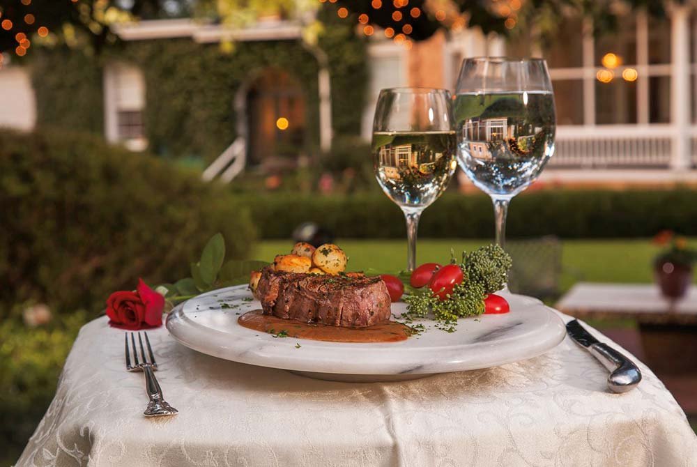 A plate with steak, potatoes, broccoli, and tomatoes sits on a table with a rose, two glasses of water, cutlery, and a white tablecloth, set outdoors in a garden setting—perfect for enjoying after exploring the many things to do in Latta and Florence SC.