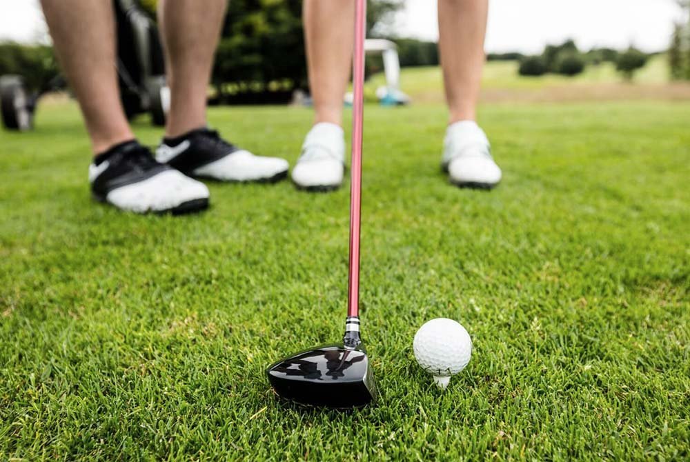 A close-up of a golf club and ball on grass, with two people standing nearby wearing athletic shoes, ready for a shot on a golf course—perfect for those looking for things to do in Latta and Florence SC.
