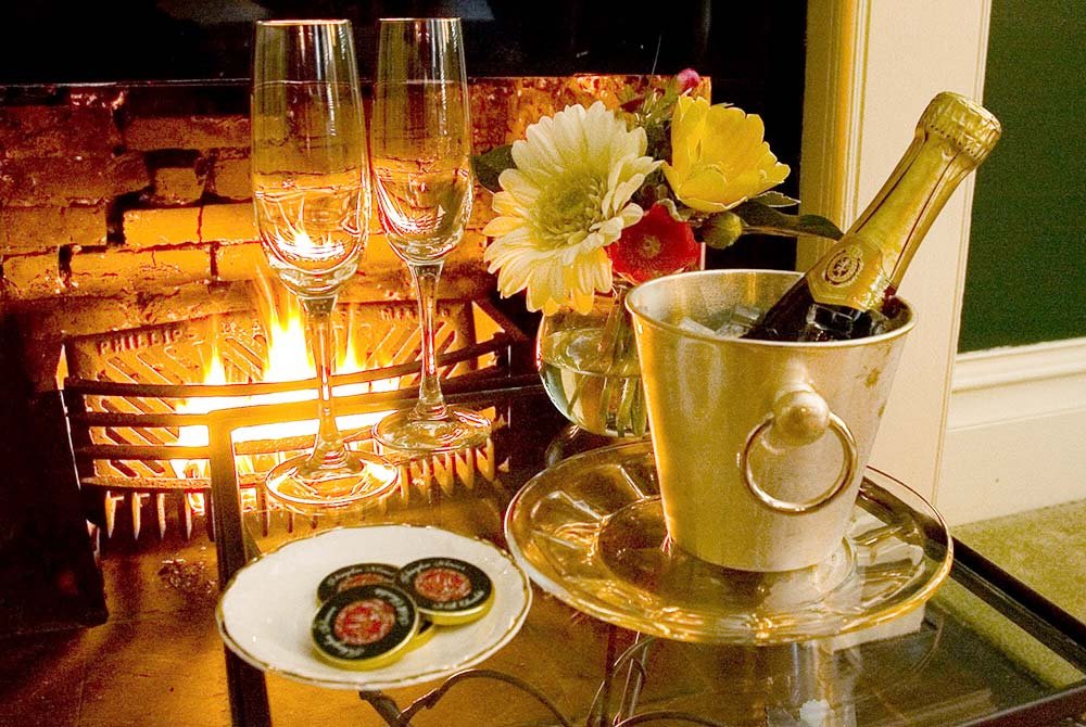 Two champagne glasses, a champagne bottle in an ice bucket, flowers, and a plate with bottle caps sit on a table in front of a lit fireplace—setting the scene for an unforgettable evening curated by exclusive South Carolina vacation packages.