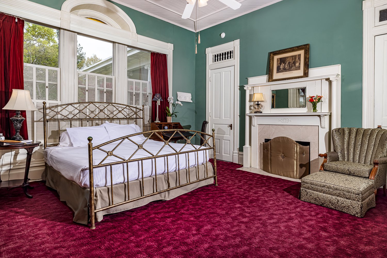 A vintage bedroom with a metal bed, green walls, red carpet, fireplace, armchair, side tables with lamps, and large windows with red curtains and white shutters—perfect for those seeking unique lodging in Florence SC.