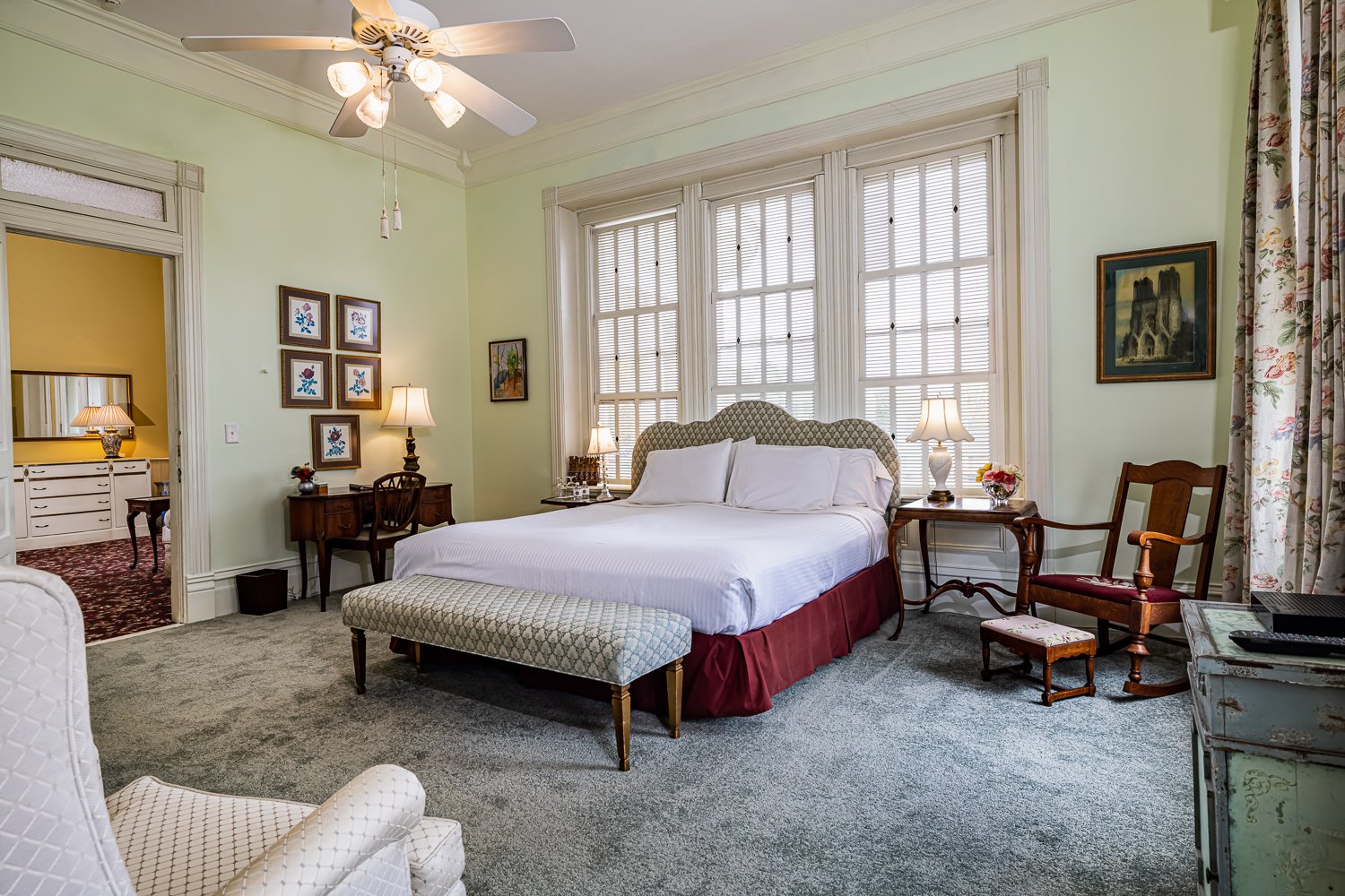 A cozy bedroom perfect for lodging in Florence SC, featuring a large bed, patterned bench, two nightstands with lamps, a wooden rocker, artwork on light green walls, and a ceiling fan above. Sunlight streams in through tall windows with blinds.