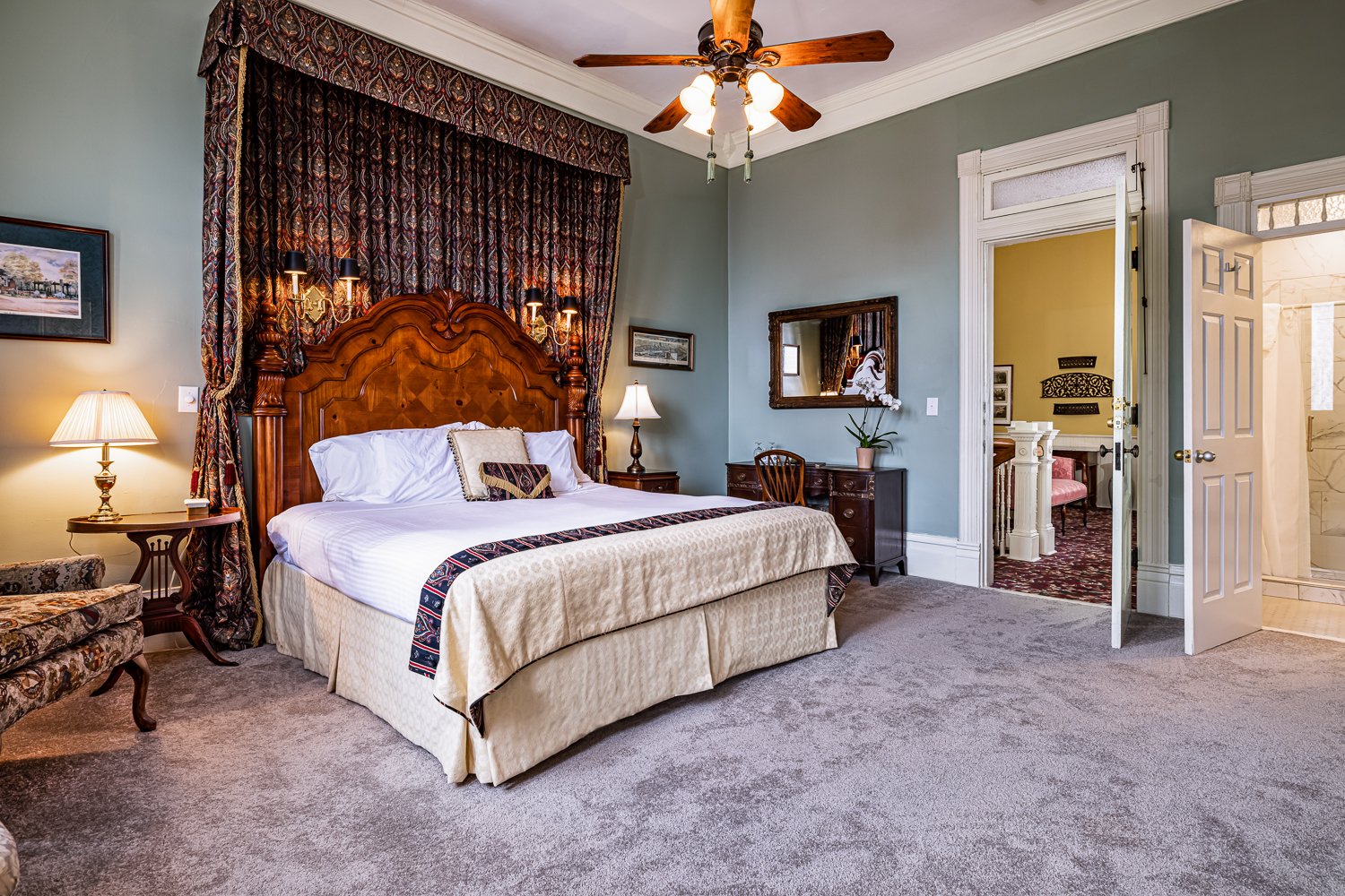 Elegant bedroom with a large wooden headboard, canopied bed, nightstands with lamps, vintage furniture, a mirror, and an open door leading to a bright room and bathroom—perfect lodging in Florence SC. Ceiling fan above.