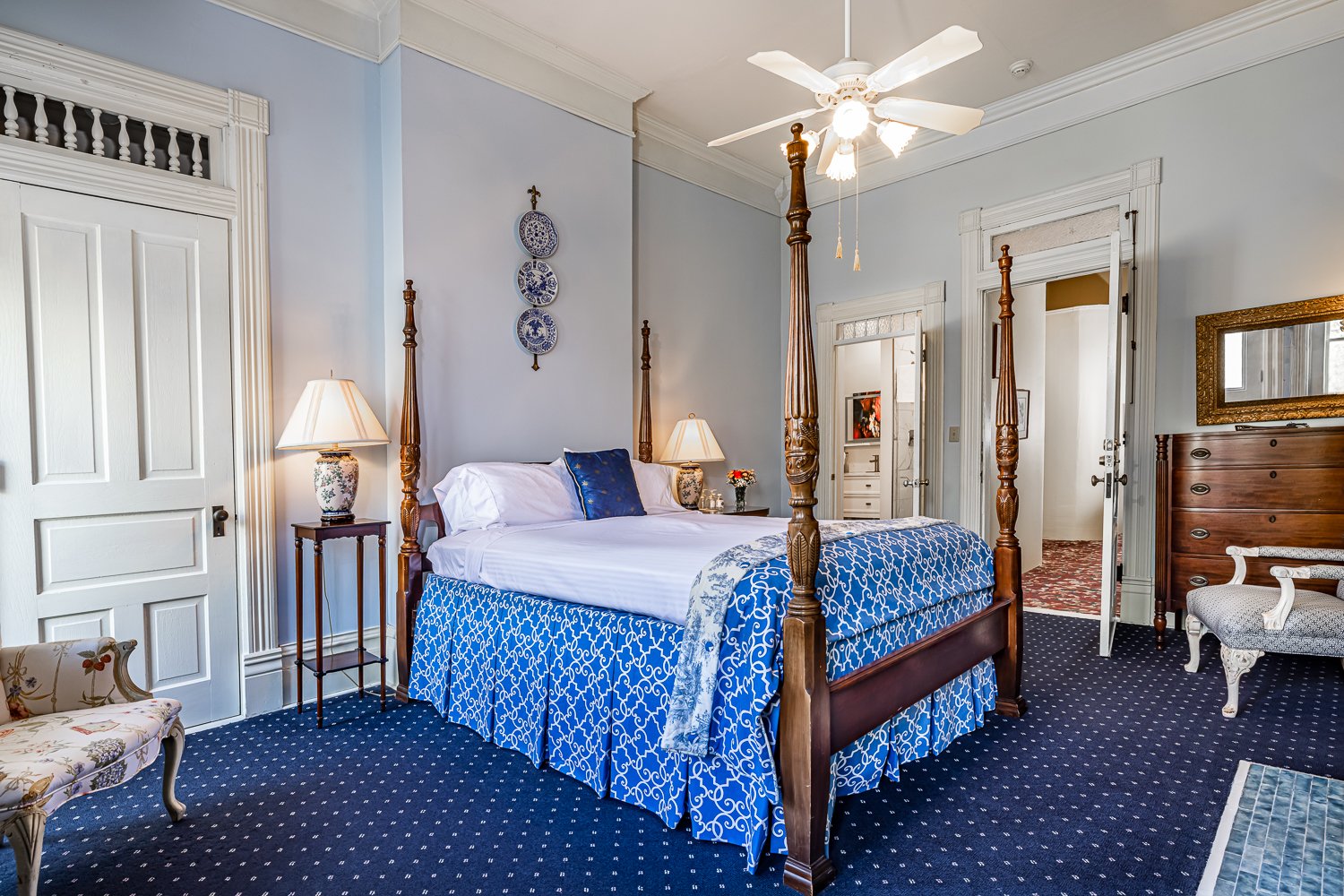 A traditional bedroom with a four-poster bed, blue and white bedding, two lamps, ornate decor, a ceiling fan, dark wood dresser, and blue carpet with white dots. Light walls and framed art add elegance, making it an inviting option for those seeking classic lodging in Florence SC.