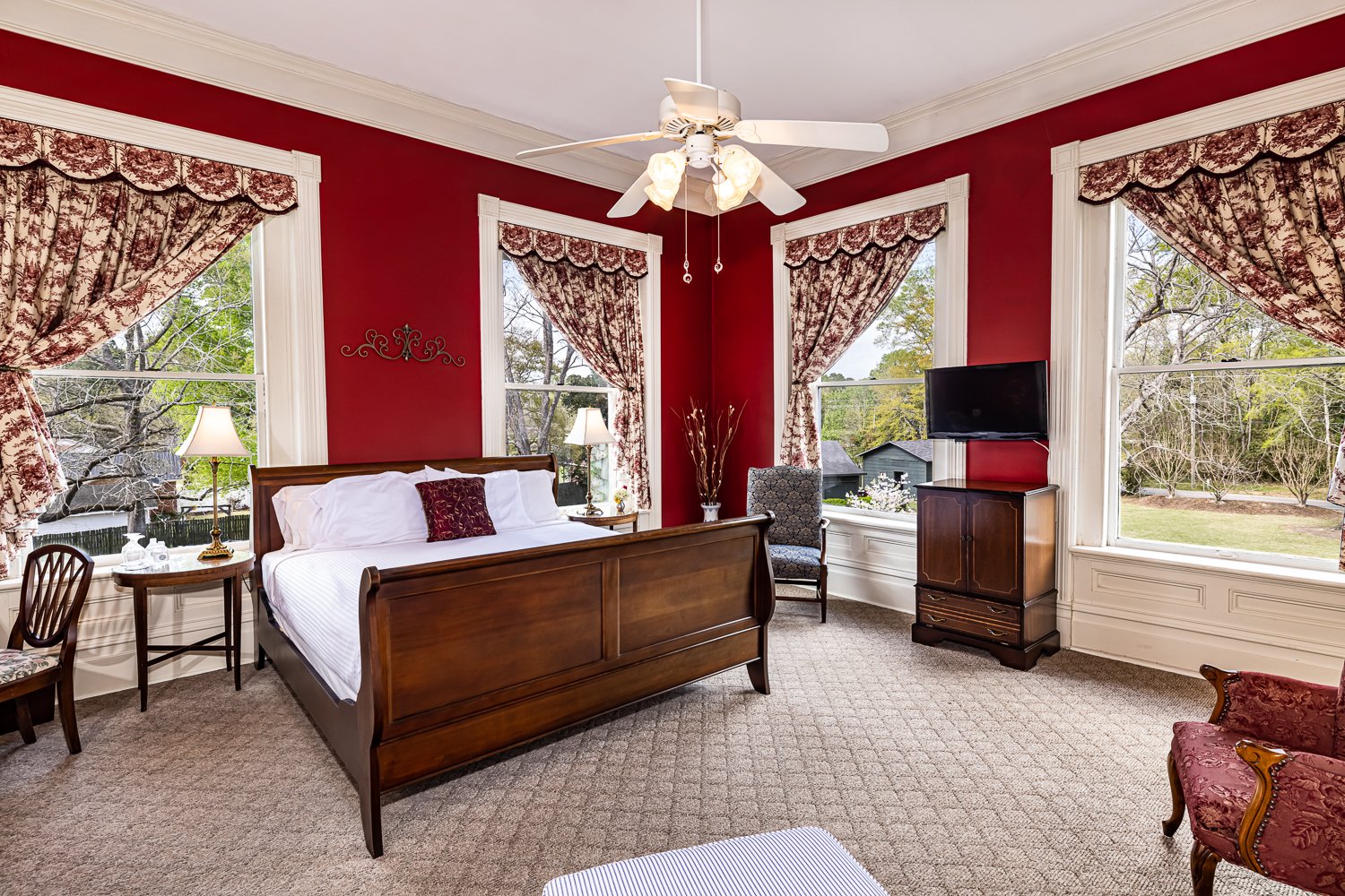 Elegant bedroom with a wooden sleigh bed, floral curtains, red walls, and large windows overlooking trees—an inviting retreat that captures the charm of classic lodging in Florence SC. A ceiling fan, lamps, TV, and vintage furnishings complete the cozy atmosphere.