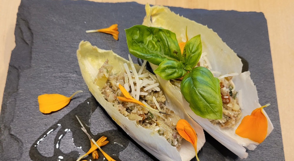 Two Belgian endive leaves filled with a savory mixture, garnished with shredded cheese, orange flower petals, and fresh basil, served on a dark slate plate with a drizzle of sauce—a refined appetizer inspired by Abingdon Manor recipes.
